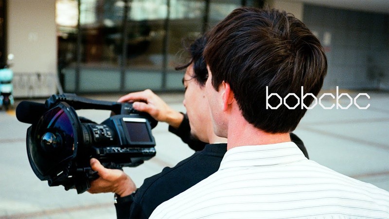 Bobby De Keyzer and Tomas Morrison looking at a video camera fitted with a wide-angle fisheye lens, with the text "bobcbc" overlaid on the right side.