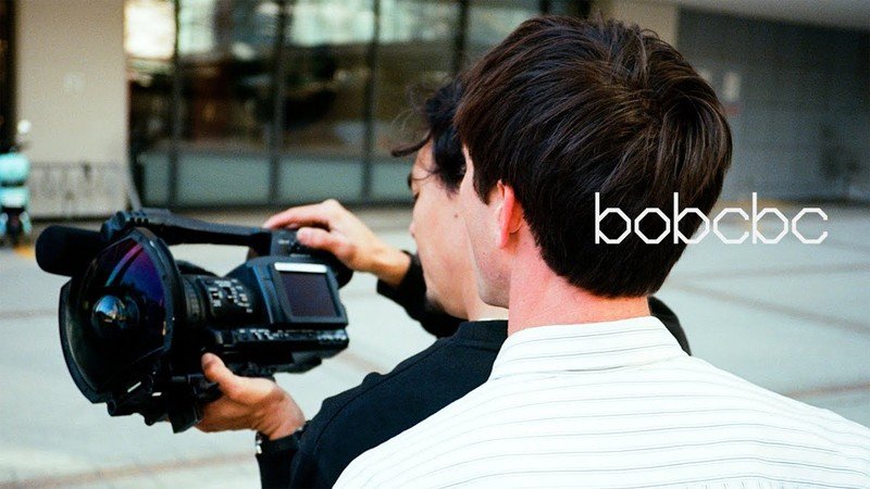 Bobby De Keyzer and Tomas Morrison looking at a video camera fitted with a wide-angle fisheye lens, with the text "bobcbc" overlaid on the right side.